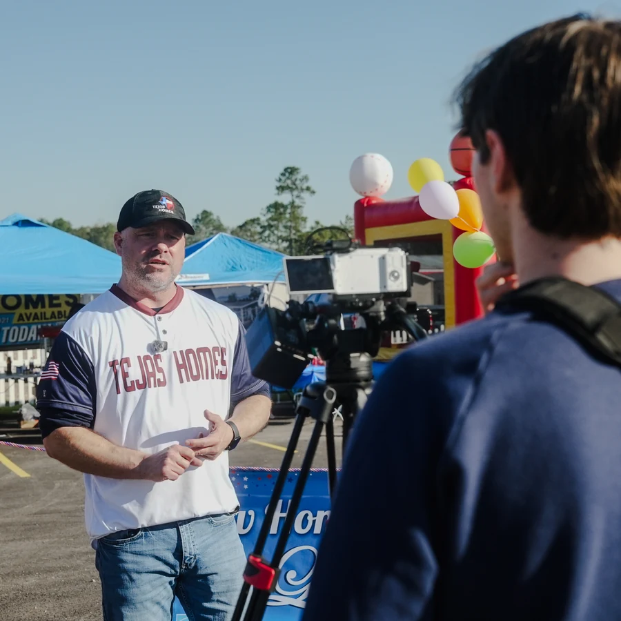 Man in a Tejas Homes jersey interviewed on camera with balloons and tents in Conroe, TX